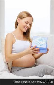 pregnancy, motherhood and celebration concept - smiling pregnant woman sitting on sofa and opening blue gift box
