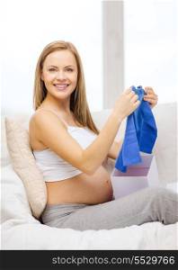 pregnancy, motherhood and celebration concept - smiling pregnant woman sitting on sofa and opening gift box with blue cardigan
