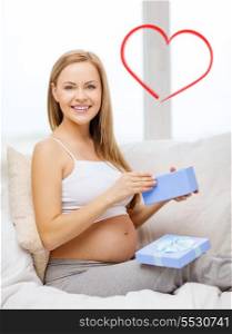 pregnancy, motherhood and celebration concept - smiling pregnant woman sitting on sofa and opening blue gift box