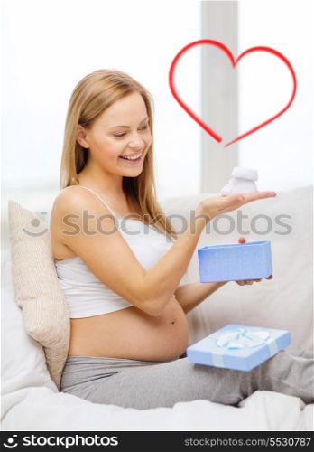 pregnancy, motherhood and celebration concept - smiling pregnant woman sitting on sofa with gift box and baby bootees