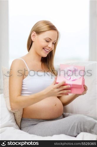 pregnancy, motherhood and celebration concept - smiling pregnant woman sitting on sofa and opening gift box