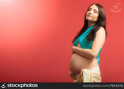 Pregnancy, motherhood and happiness concept. Attractive pregnant woman in scarf touching her belly on red background