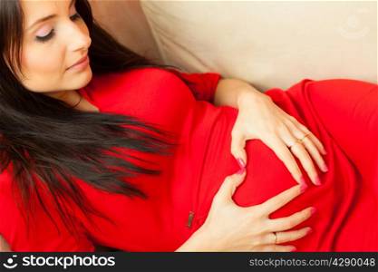 Pregnancy, motherhood and happiness concept. Beautiful stylish elegant pregnant woman in red dress relaxing on sofa, making heart shape with hands over belly, symbol of love