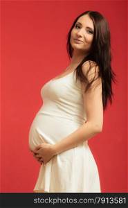 Pregnancy, motherhood and happiness concept. Elegant pregnant woman in white dress touching her belly at home