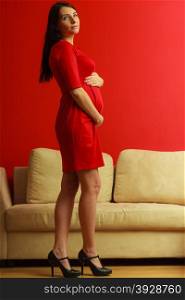 Pregnancy, motherhood and happiness concept. Full length elegant pregnant woman in short red dress touching her belly at home