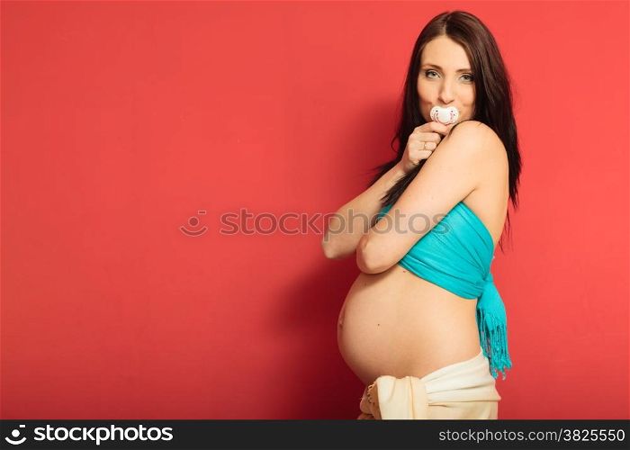 Pregnancy, motherhood and happiness concept. Funny pregnant woman holding dummy for the unborn baby in mouth on red background
