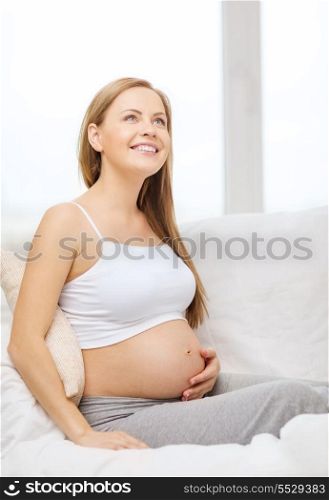 pregnancy, motherhood and happiness concept - happy pregnant woman sitting on sofa and touching her belly and dreaming