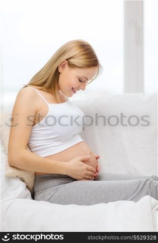 pregnancy, motherhood and happiness concept - happy pregnant woman sitting on sofa and touching her belly