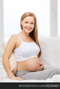 pregnancy, motherhood and happiness concept - happy pregnant woman sitting on sofa and touching her belly