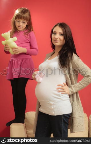 Pregnancy, motherhood and happiness concept. pregnant woman holding small shoes boots for the unborn baby, little girl playing in the backgroynd