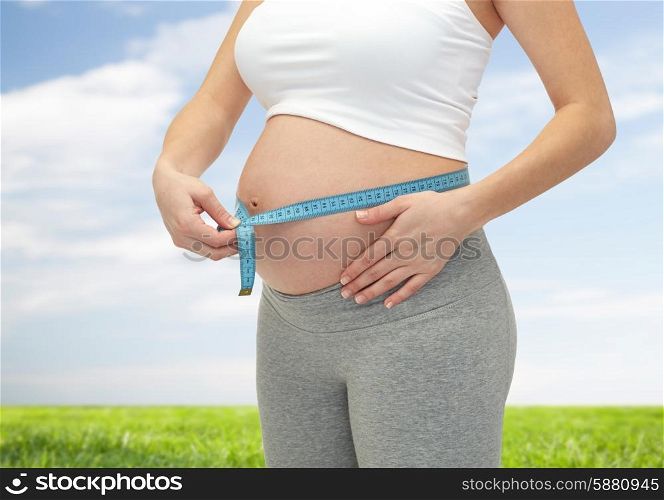 pregnancy, motherhood, control, people and expectation concept - close up of happy pregnant woman measuring her bare tummy over blue sky and grass background