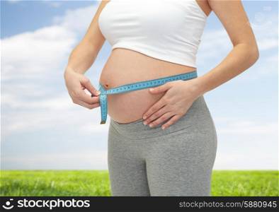pregnancy, motherhood, control, people and expectation concept - close up of happy pregnant woman measuring her bare tummy over blue sky and grass background