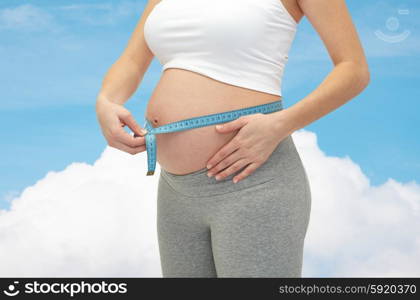pregnancy, motherhood, control, people and expectation concept - close up of happy pregnant woman measuring her bare tummy over blue sky and cloud background