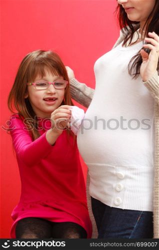 Pregnancy, motherhood, family concept. Pregnant woman with her daughter holding little white baby socks and touching her belly.. Pregnant woman with toddler girl holding baby shoes