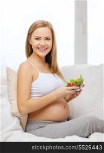 pregnancy, motherhood, healthcare, food and happiness concept - happy pregnant woman sitting on sofa with bowl of salad