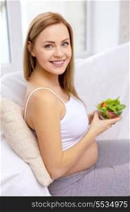 pregnancy, motherhood, healthcare, food and happiness concept - happy pregnant woman sitting on sofa with bowl of salad