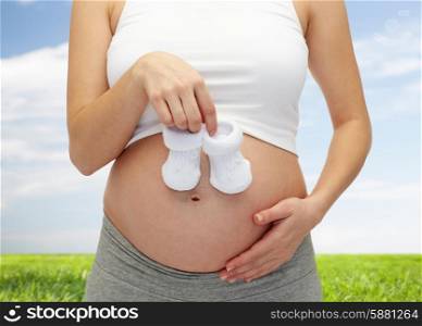 pregnancy, motherhood, people and expectation concept - close up of happy pregnant woman touching her bare tummy and holding babys bootees over blue sky and grass background