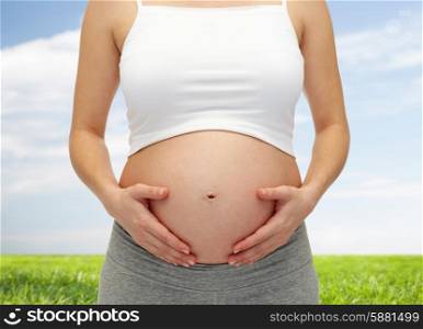 pregnancy, motherhood, people and expectation concept - close up of pregnant woman touching her bare tummy over blue sky and grass background
