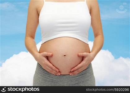 pregnancy, motherhood, people and expectation concept - close up of pregnant woman touching her bare tummy over blue sky and cloud background