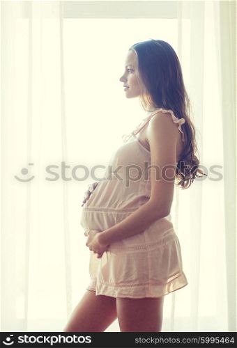 pregnancy, motherhood, people and expectation concept - happy pregnant woman in chemise near window at home