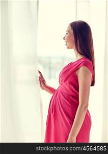 pregnancy, motherhood, people and expectation concept - happy pregnant woman with big tummy looking through window at home