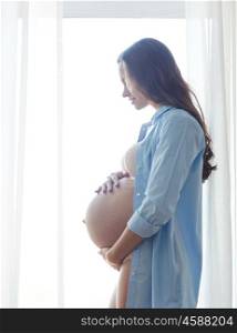 pregnancy, motherhood, people and expectation concept - happy pregnant woman with big bare tummy near window at home