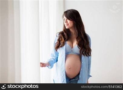 pregnancy, motherhood, people and expectation concept - happy pregnant woman with big bare tummy looking through window at home. happy pregnant woman with big tummy at home