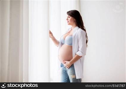 pregnancy, motherhood, people and expectation concept - happy pregnant woman with big bare tummy looking through window at home. happy pregnant woman with big tummy at home