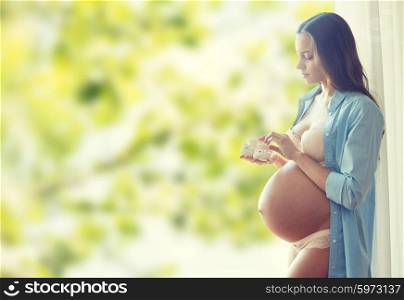 pregnancy, motherhood, people and expectation concept - happy pregnant woman with big bare tummy holding little baby booties at home over green natural background
