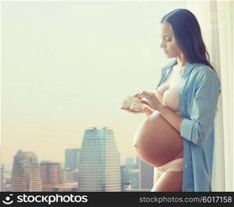 pregnancy, motherhood, people and expectation concept - happy pregnant woman with big bare tummy holding little baby booties at home over city background