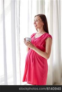 pregnancy, motherhood, people and expectation concept - happy pregnant woman with cup drinking and looking through window at home
