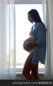 pregnancy, motherhood, people and expectation concept - silhouette of happy pregnant woman with big bare tummy near window at home