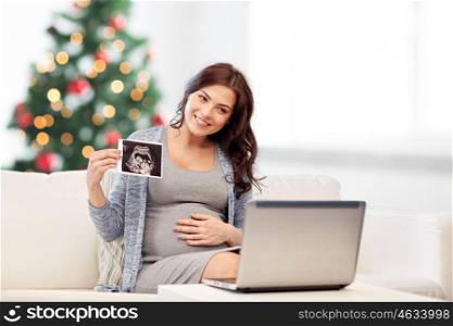 pregnancy, motherhood, people and holidays concept - happy pregnant woman with laptop computer having video call and showing ultrasound image over christmas tree background