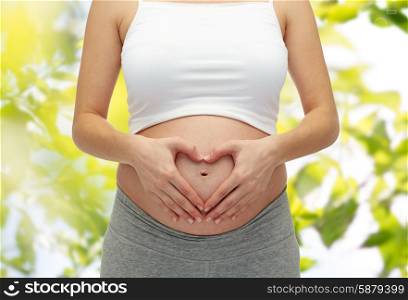 pregnancy, motherhood, people, love and expectation concept - close up of happy pregnant woman making heart shape hand sign on her bare tummy over green tree leavers background