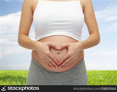 pregnancy, motherhood, people, love and expectation concept - close up of happy pregnant woman making heart shape hand sign on her bare tummy over blue sky and grass background