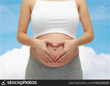 pregnancy, motherhood, people, love and expectation concept - close up of happy pregnant woman making heart shape hand sign on her bare tummy over blue sky and cloud background