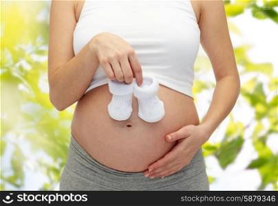 pregnancy, motherhood, people, summer and expectation concept - close up of happy pregnant woman touching her bare tummy and holding babys bootees over green tree leavers background