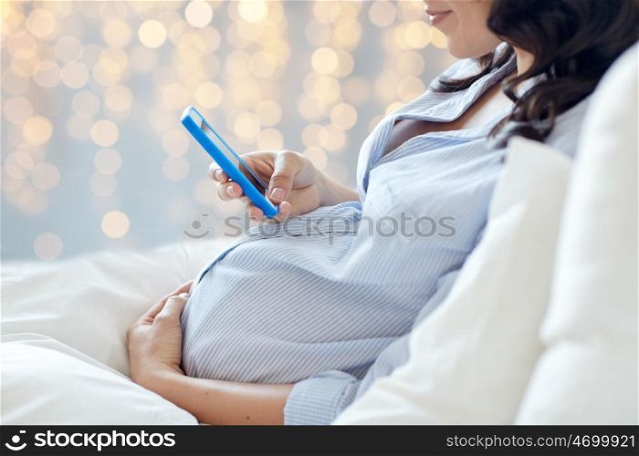 pregnancy, motherhood, technology, people and expectation concept - close up of pregnant woman with smartphone in bed over holidays lights background