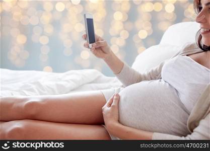 pregnancy, motherhood, technology, people and expectation concept - close up of pregnant woman with smartphone in bed over holidays lights background