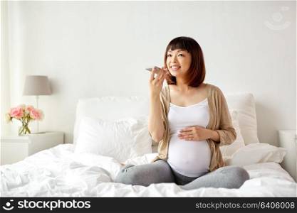 pregnancy, motherhood, technology, people and expectation concept - happy pregnant asian woman using voice command recorder or calling on smartphone at home. pregnant woman using voice recorder on smartphone