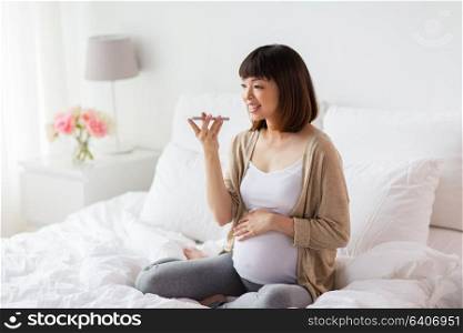 pregnancy, motherhood, technology, people and expectation concept - happy pregnant asian woman using voice command recorder or calling on smartphone at home. pregnant woman using voice recorder on smartphone
