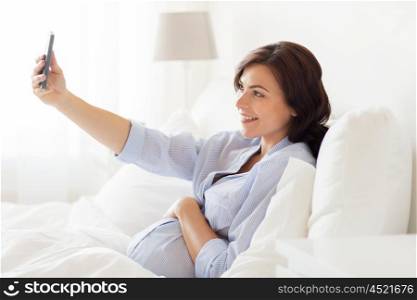 pregnancy, motherhood, technology, people and expectation concept - happy pregnant woman with smartphone taking selfie in bed at home