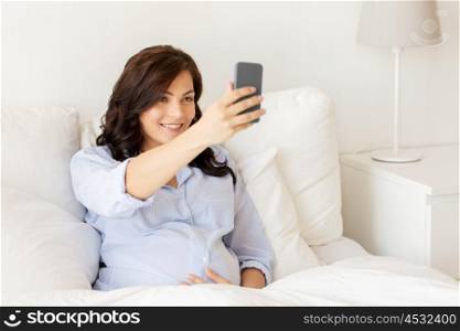 pregnancy, motherhood, technology, people and expectation concept - happy pregnant woman with smartphone taking selfie in bed at home