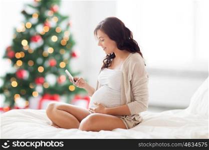 pregnancy, motherhood, technology, people and expectation concept - happy pregnant woman with smartphone in bed over christmas tree background