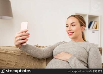pregnancy, motherhood, technology, people and expectation concept - happy pregnant woman with smartphone taking selfie at home. pregnant woman taking smartphone selfie at home