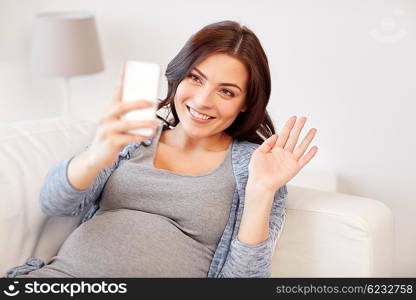 pregnancy, motherhood, technology, people and expectation concept - happy pregnant woman with smartphone having video call and waving hand at home