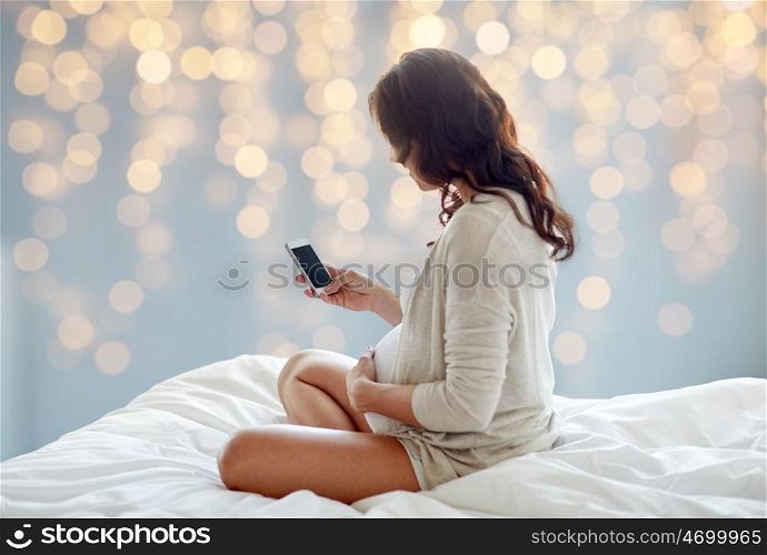 pregnancy, motherhood, technology, people and expectation concept - pregnant woman with smartphone in bed over holidays lights background
