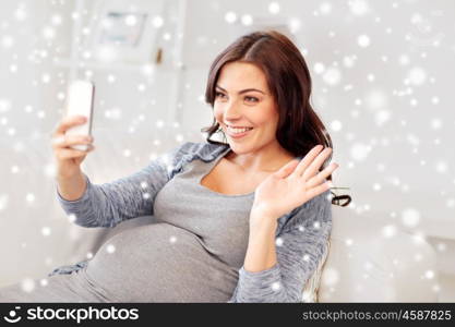 pregnancy, motherhood, technology, people and winter concept - happy pregnant woman with smartphone having video call and waving hand at home over snow