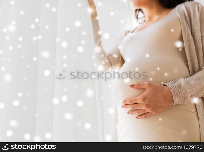 pregnancy, motherhood, winter, people and expectation concept - close up of happy pregnant woman with big belly looking to window over snow