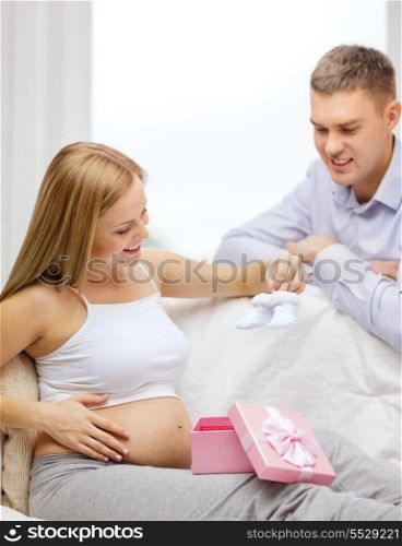 pregnancy, parenthood and celebration concept - happy family expectin child sitting on sofa with gift box and baby bootees